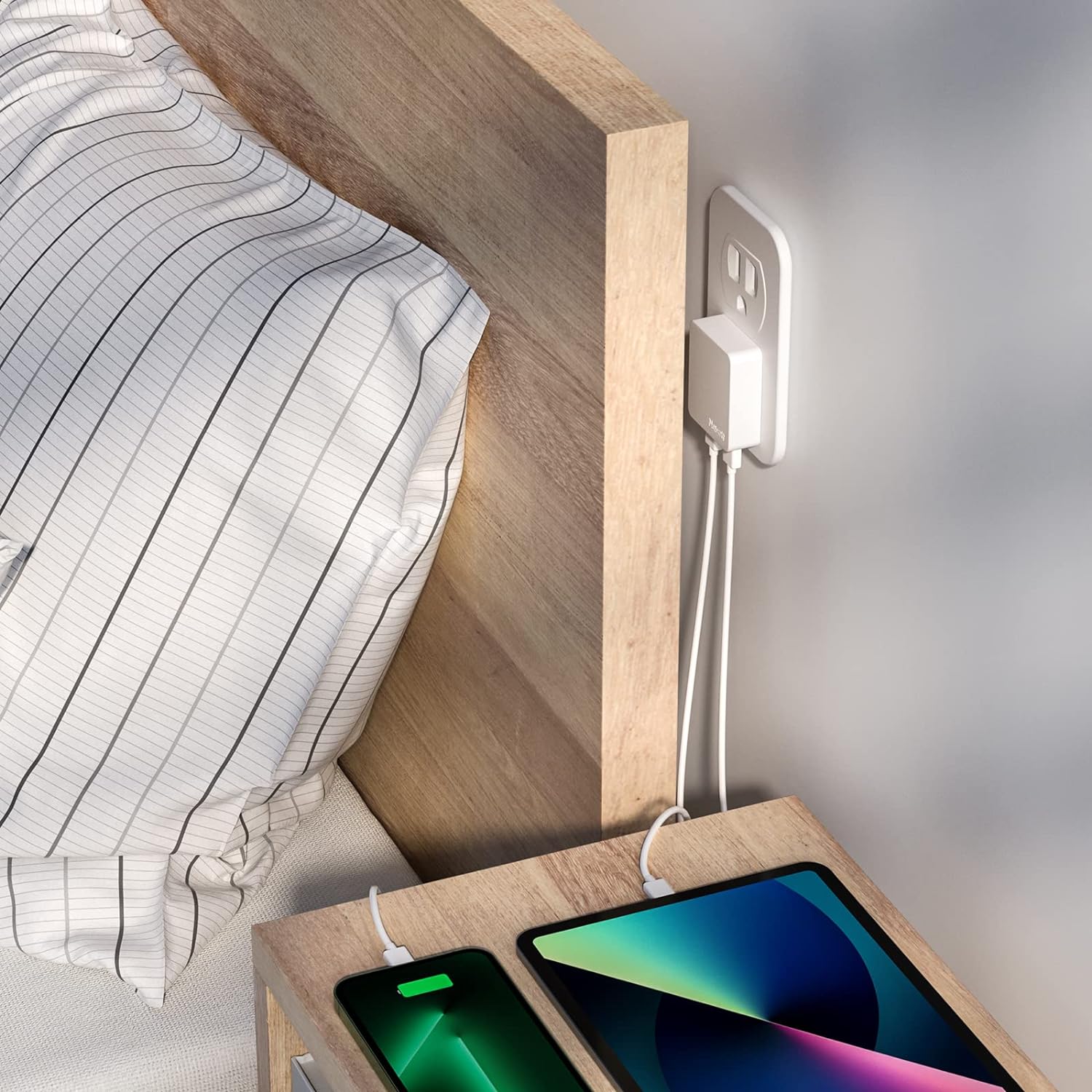 Two smartphones on a wooden nightstand with a power adapter plugged into a wall socket.