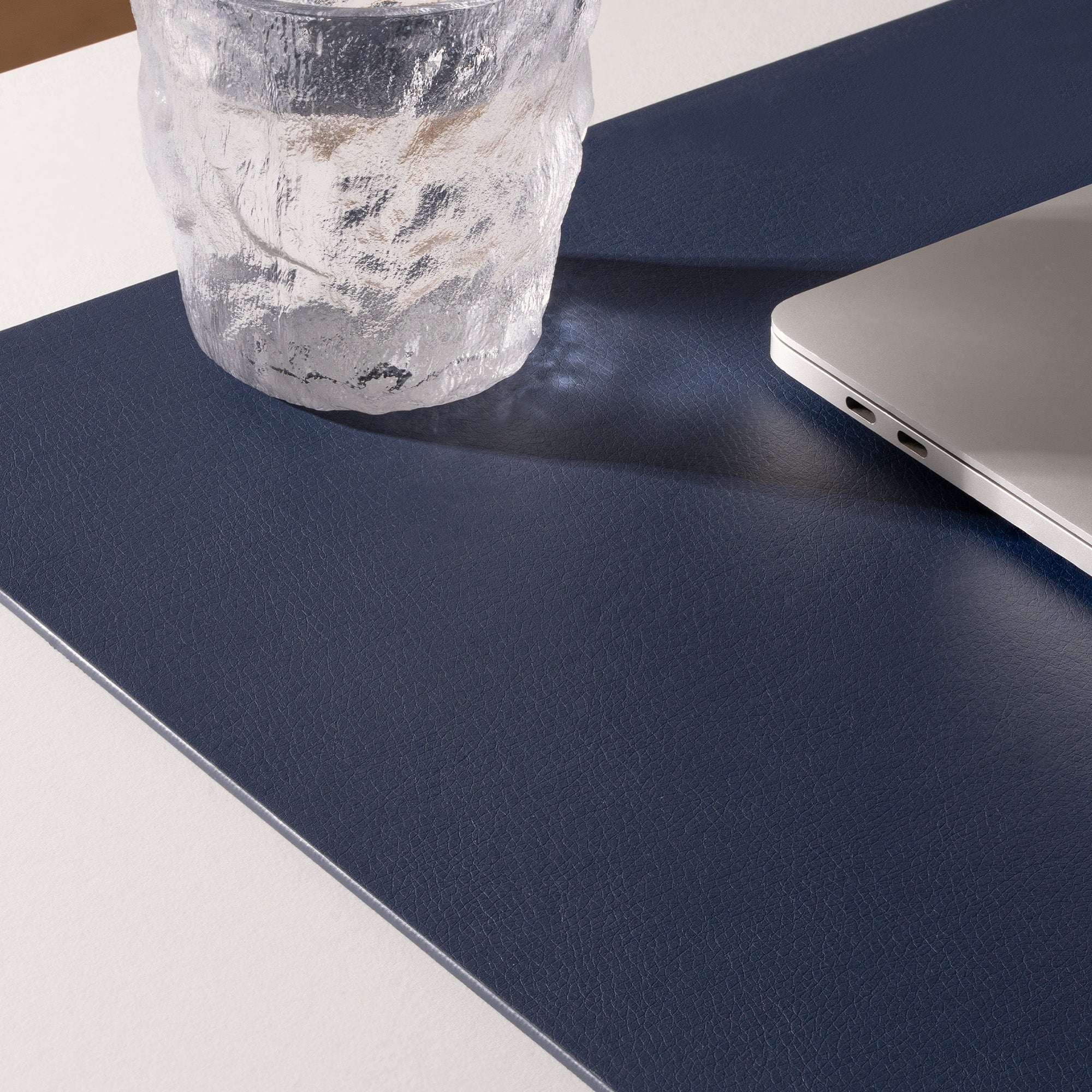 Navy blue desk pad with a laptop and clear vase on a light gray surface