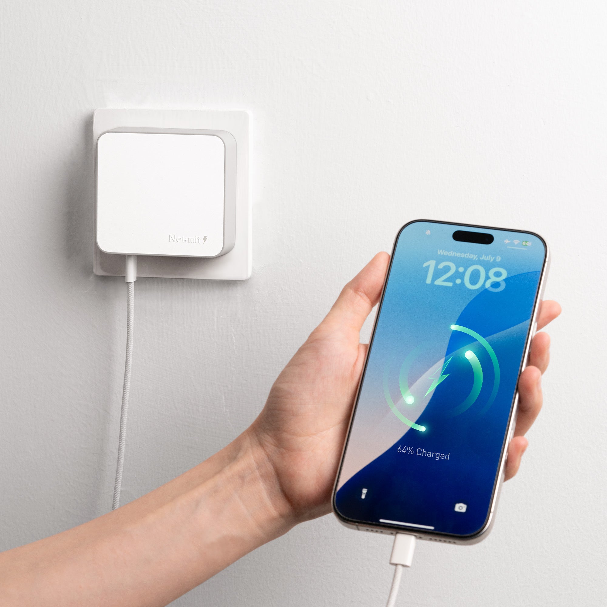 Person holding a smartphone with a charging cable connected to a wall-mounted device on a light gray background