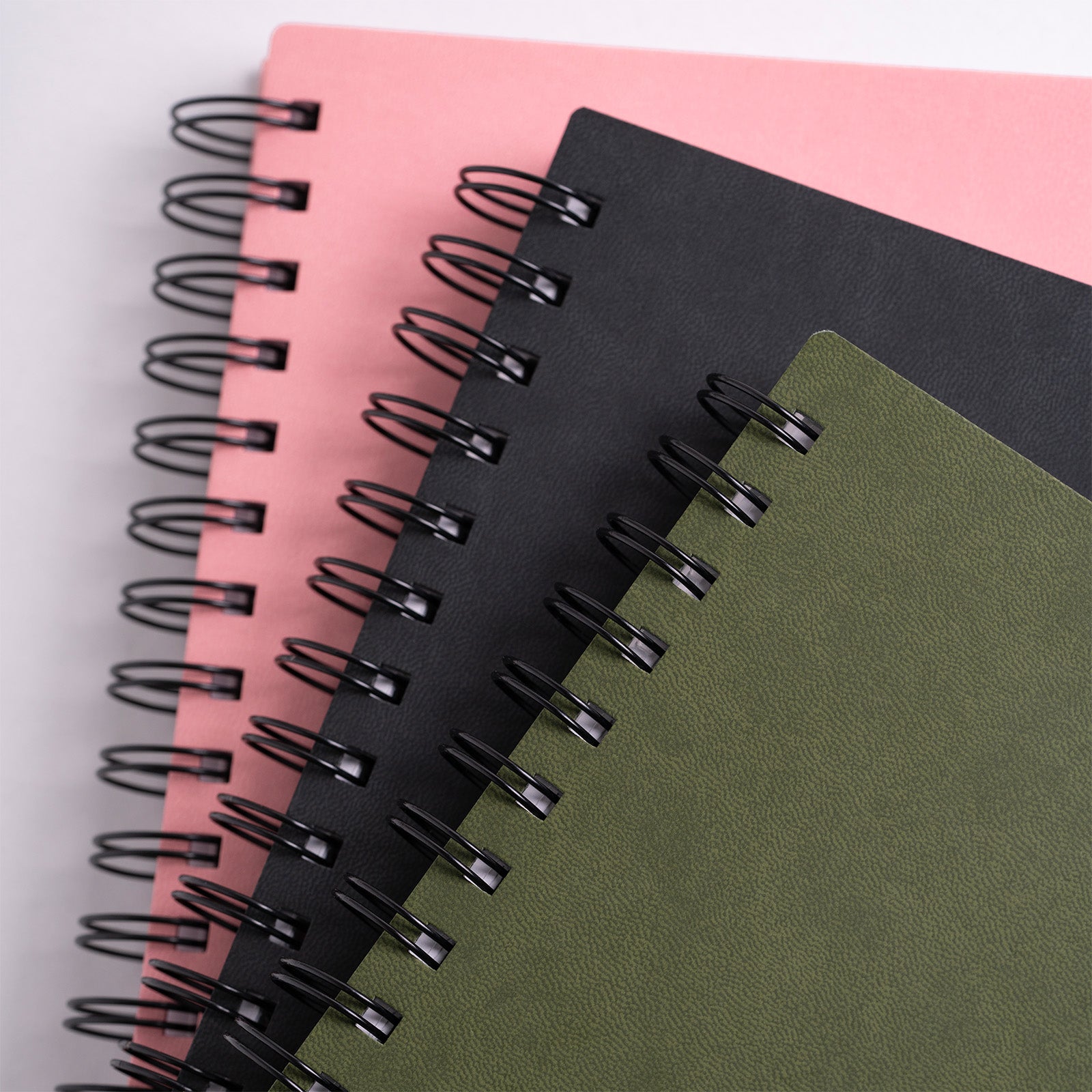 Three spiral-bound notebooks in pink, black, and green on a light gray background