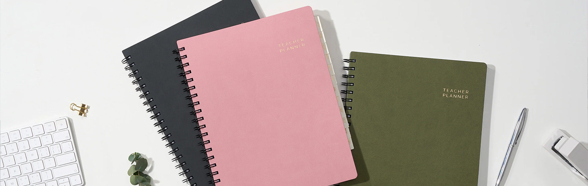Four spiral-bound planners in black, pink, and green on a white surface with office supplies.