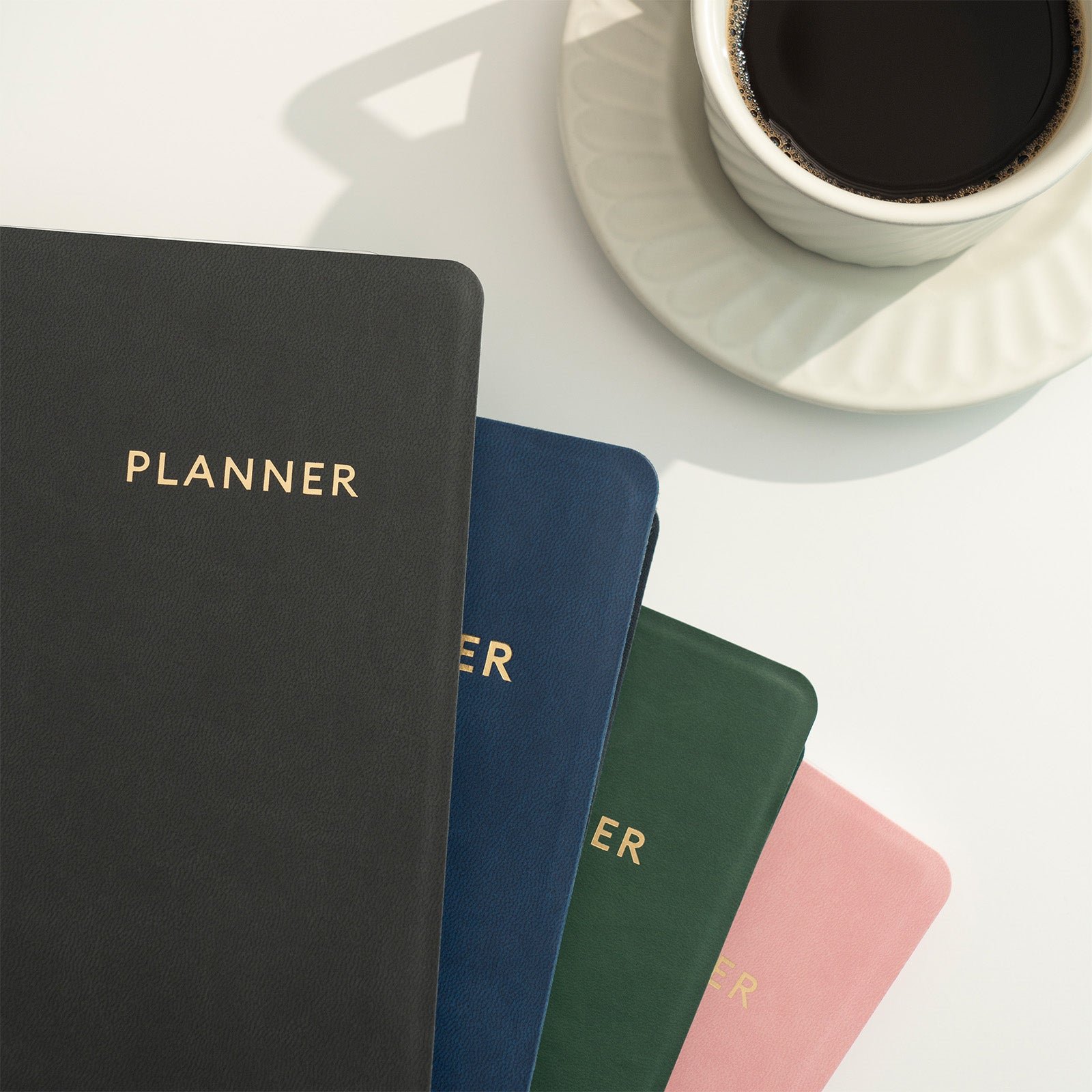Four planners in black, blue, green, and pink with 'PLANNER' text on a white surface with a cup of coffee.