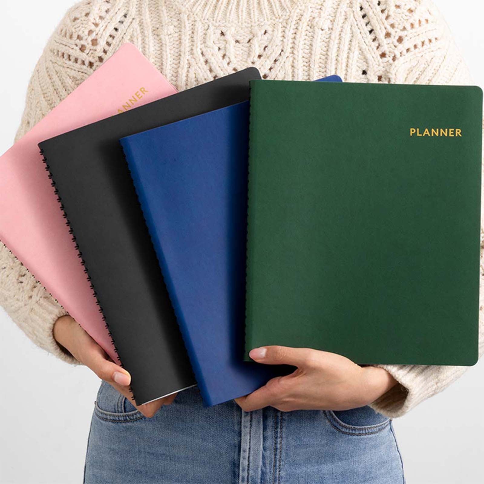 Four planners in different colors held by a person against a neutral background