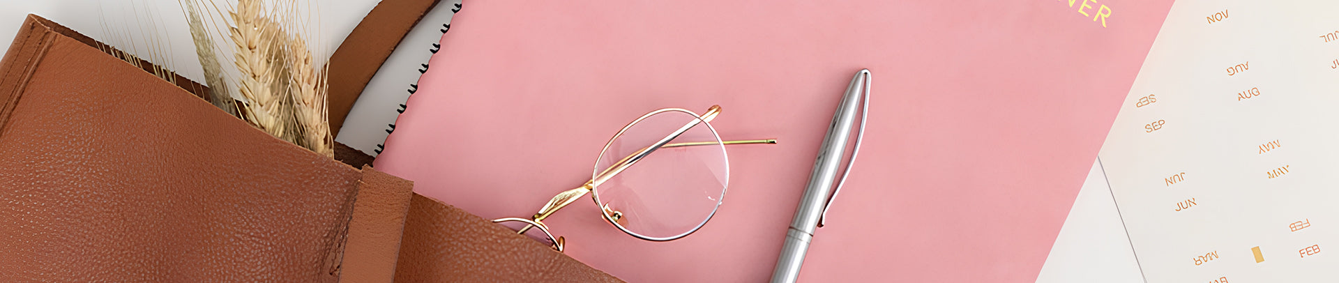 Brown notebook, pink notepad, glasses, and pen on a white surface