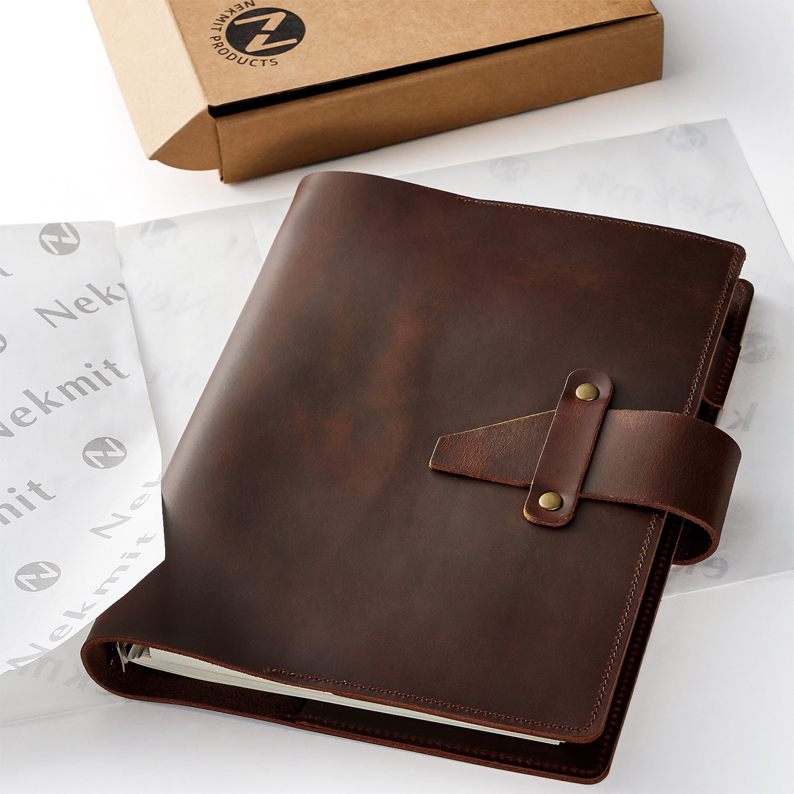 Brown leather notebook with strap closure on a white surface with 'Nekmit' branding.