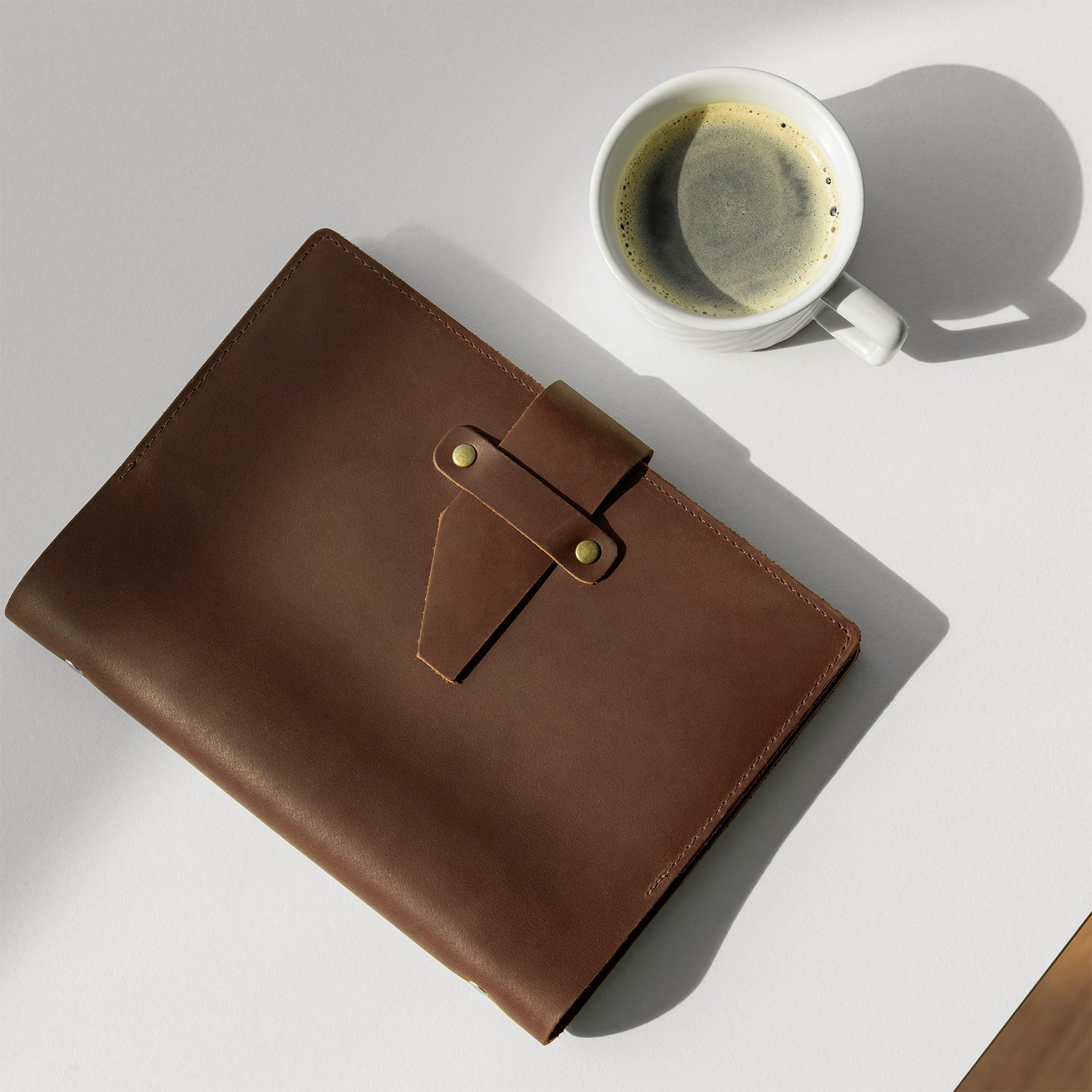 Brown leather notebook and white mug with coffee on a light gray surface