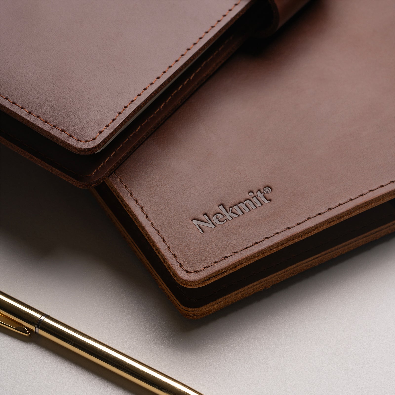 Brown leather notebook with 'Nekmit' branding and a gold pen on a light surface