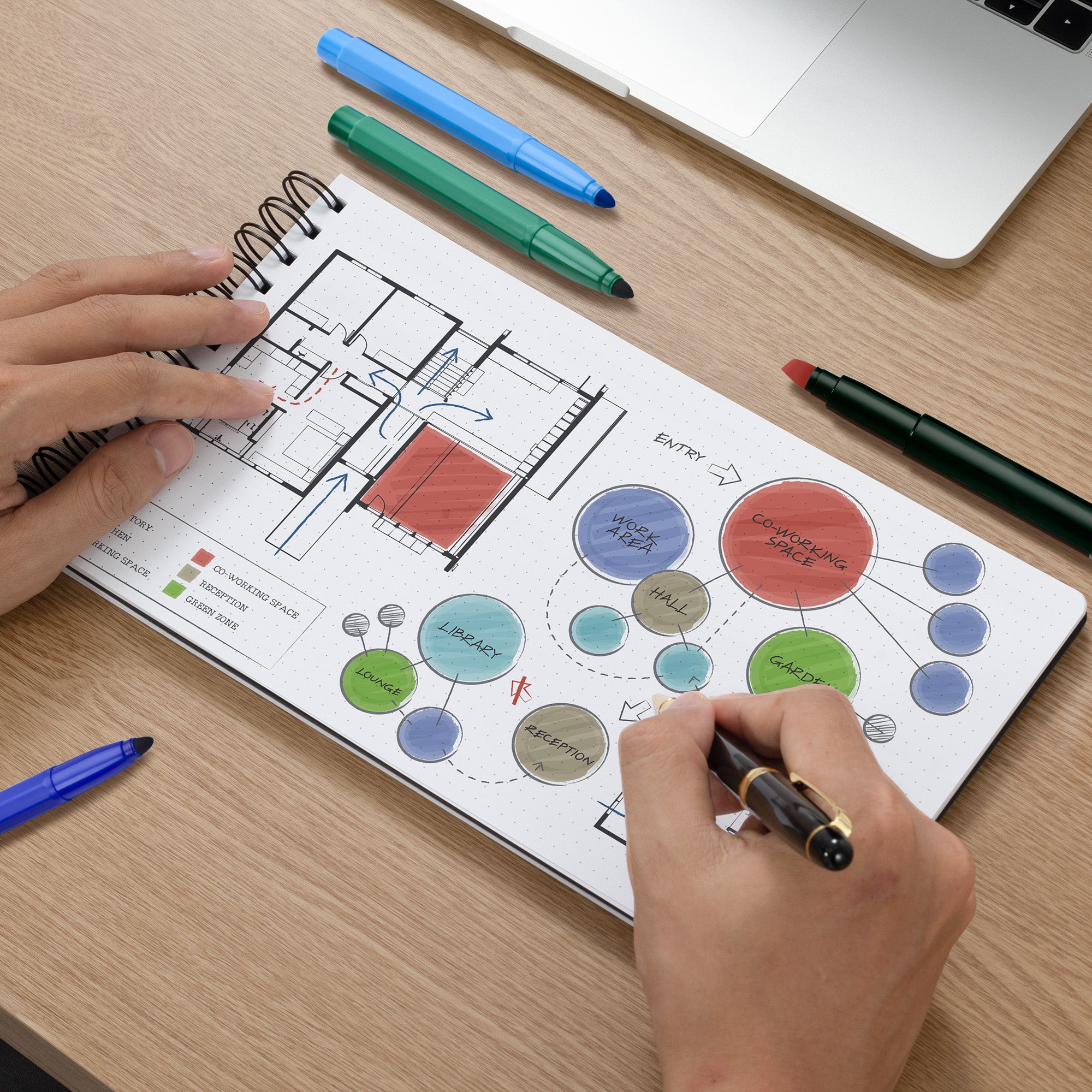 Person drawing a floor plan and diagram on a notebook with markers on a wooden desk.