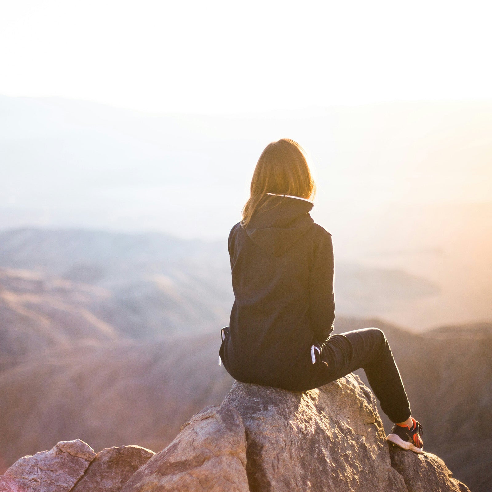 Person sitting on a rocky outcrop overlooking a vast landscape with the sun setting.